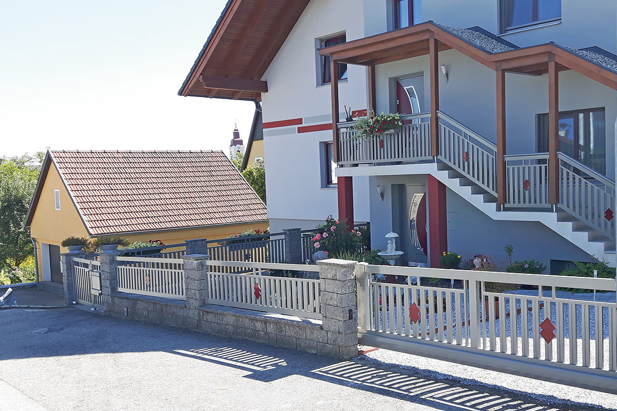 Grauer Gartenzaun mit roten Elementen mit traditionellen Haus im Hintergrund